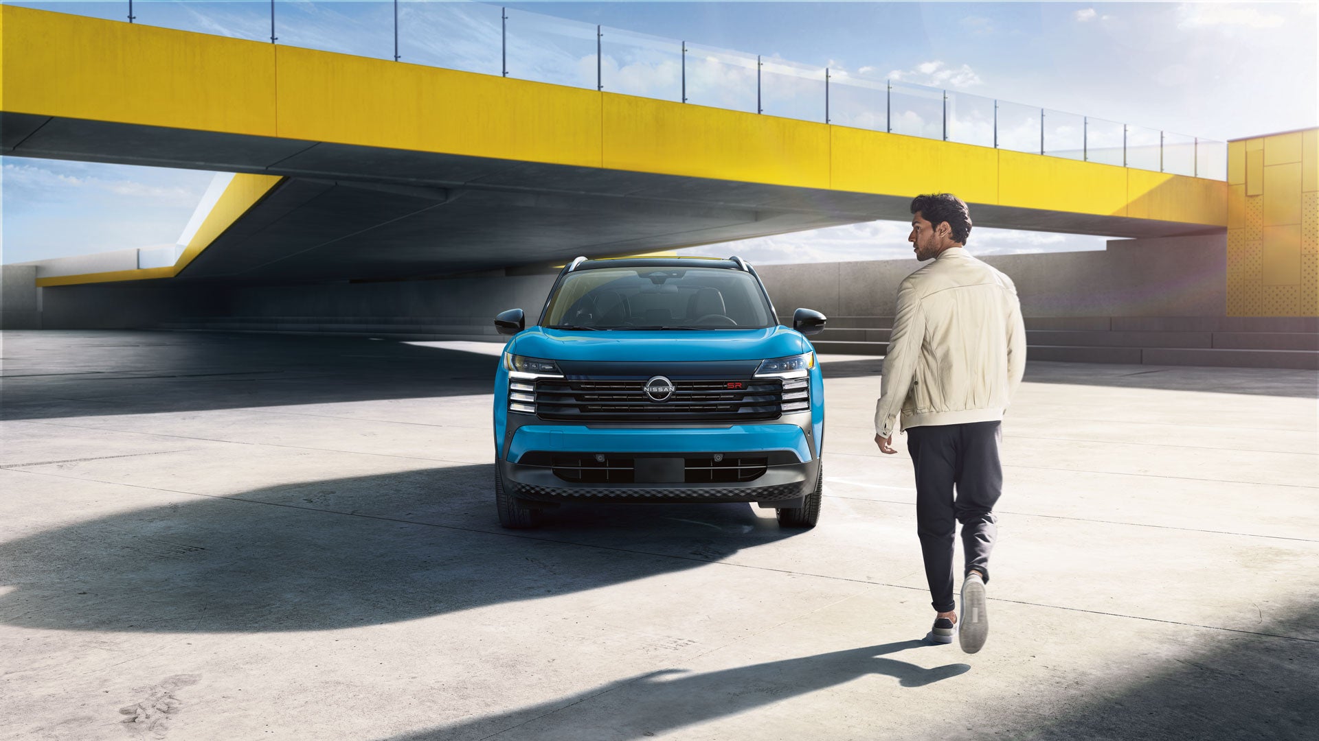 A man in a cream jacket and dark pants walks away from a blue Nissan Kicks parked under a yellow overpass.