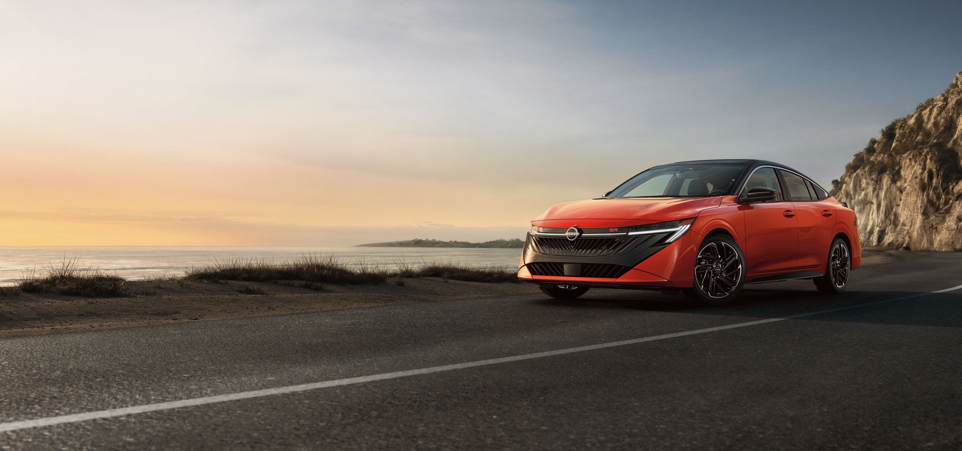 A red electric car with a black roof drives on a coastal road at sunset, with the ocean and hills in the background.