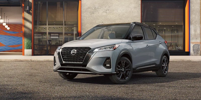 A light gray Nissan Kicks SUV with a black roof and black wheels is parked on asphalt in front of a modern storefront.