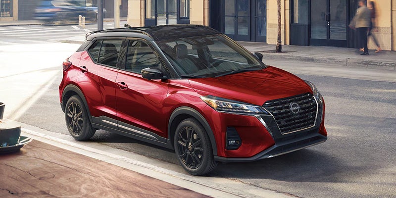 A red Nissan Kicks with a black roof parked on a city street, with people and buildings in the background.