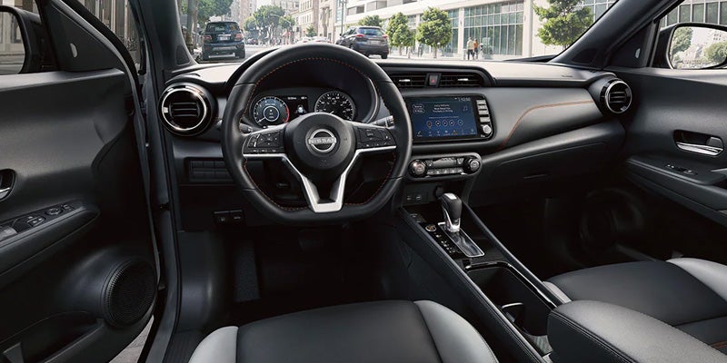 Interior shot of a Nissan Kicks dashboard and steering wheel, with black and gray seats and city street visible outside.