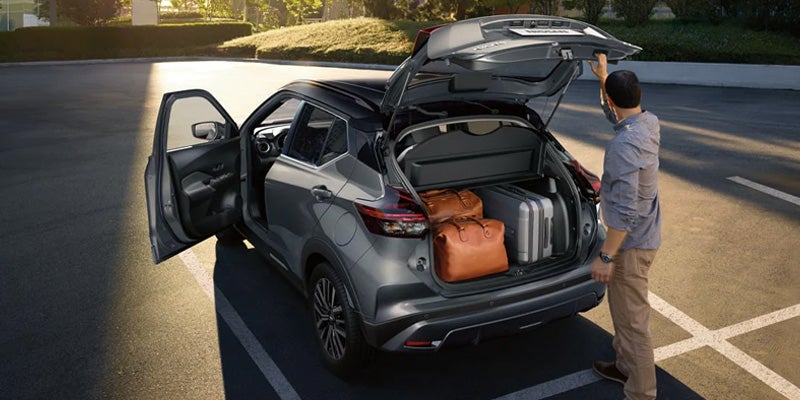 A man loads two suitcases and a bag into the open trunk of a gray Nissan Kicks in a sunny parking lot.