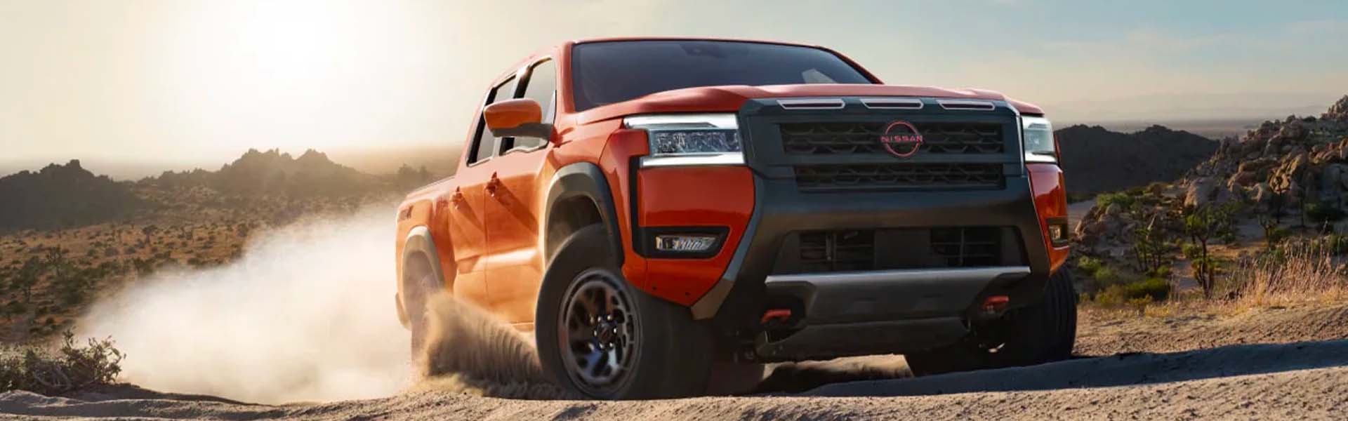 Orange Nissan Frontier pickup driving on a dusty desert road, mountains in the hazy background.