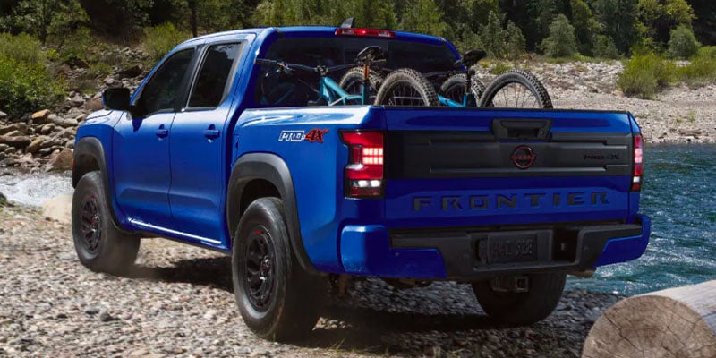 Rear view of a blue Nissan Frontier PRO-4X pickup truck with two mountain bikes in the bed, by a river.