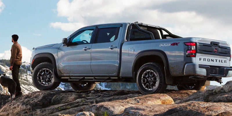 Light gray Nissan Frontier pickup truck parked on a rocky mountain ledge with a person and dog on the left.