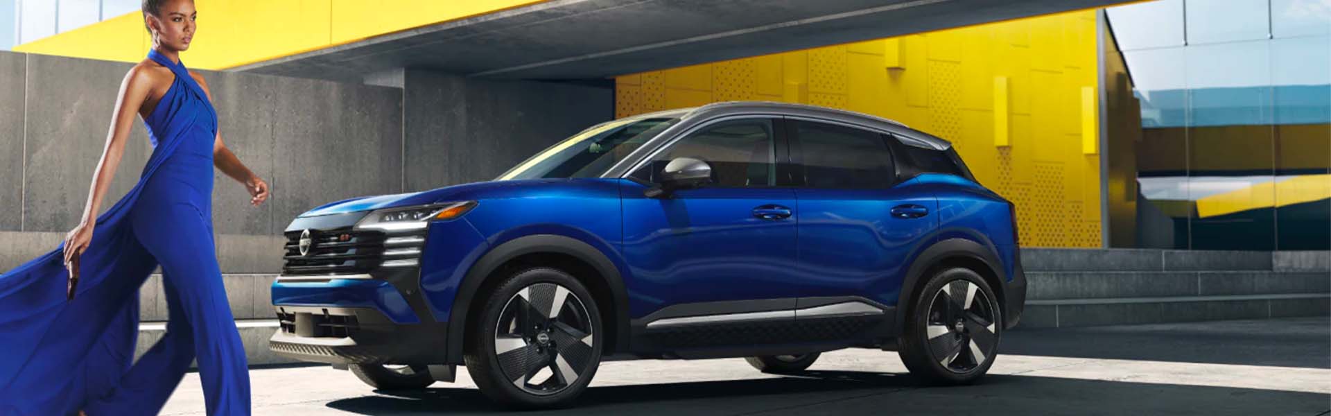 A woman in a flowing blue jumpsuit poses next to a sleek blue crossover SUV against concrete and yellow walls.