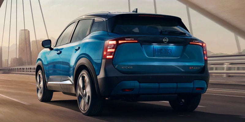 Rear view of a blue Nissan Kicks SUV driving on a bridge, with a distant city skyline in the background.