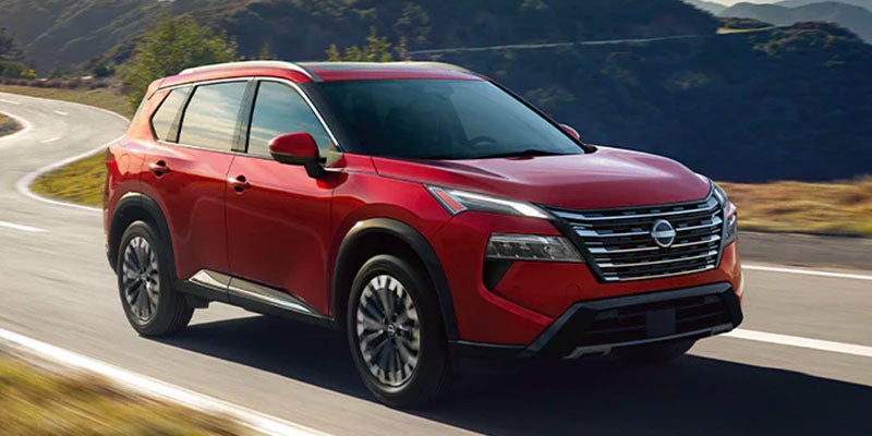 A red Nissan Rogue SUV drives on a winding road with mountains in the background.