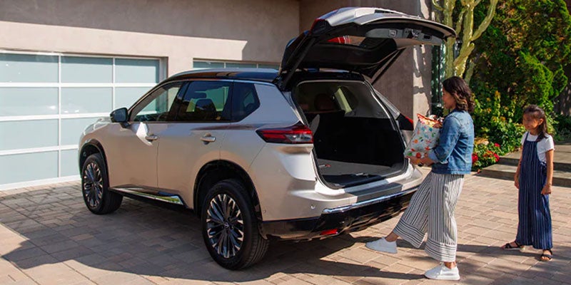 Woman and child load groceries into the open trunk of a light-colored Nissan Rogue in a driveway.