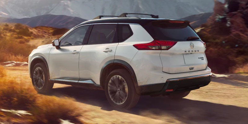 Rear right angle view of a white Nissan Rogue SUV with black roof and roof rack driving on a dirt road.