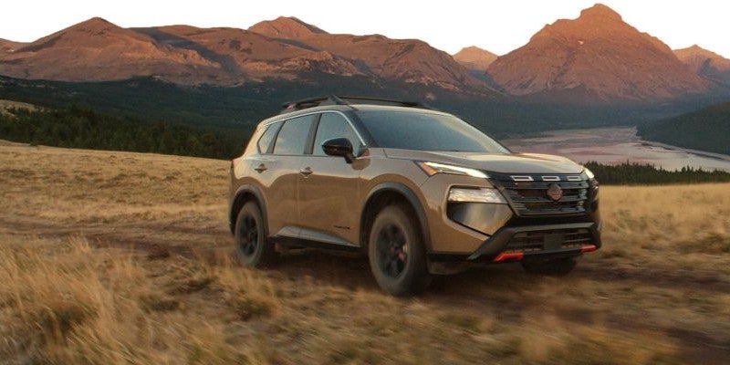 Brown SUV driving on a dirt path through golden grass with mountains and a river in the background.