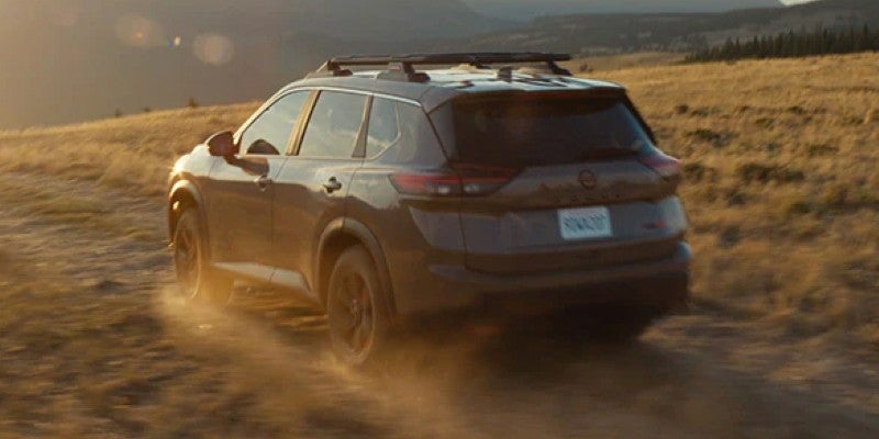 Dark grey Nissan Pathfinder SUV driving on a dirt road, kicking up dust at sunset. Hills in the golden background.