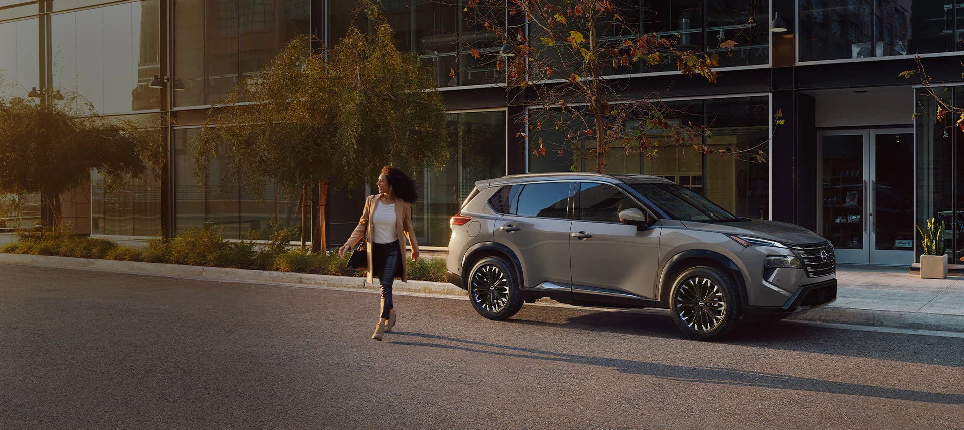 A woman walks past a gray SUV parked on a city street next to modern glass buildings.
