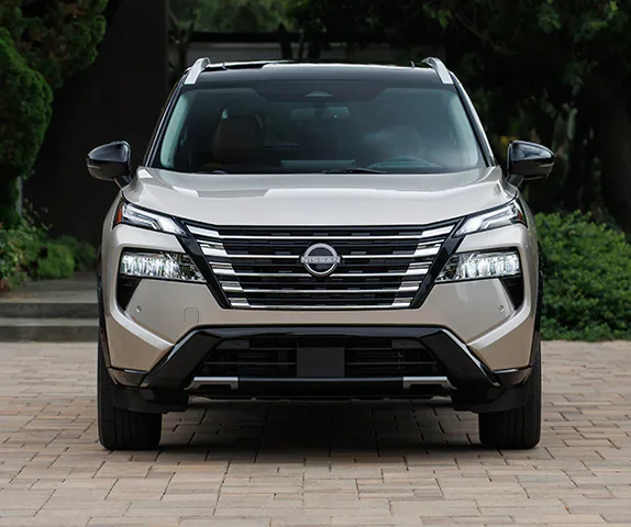 Front view of a light gold Nissan Rogue SUV parked on a paved driveway in front of a house.