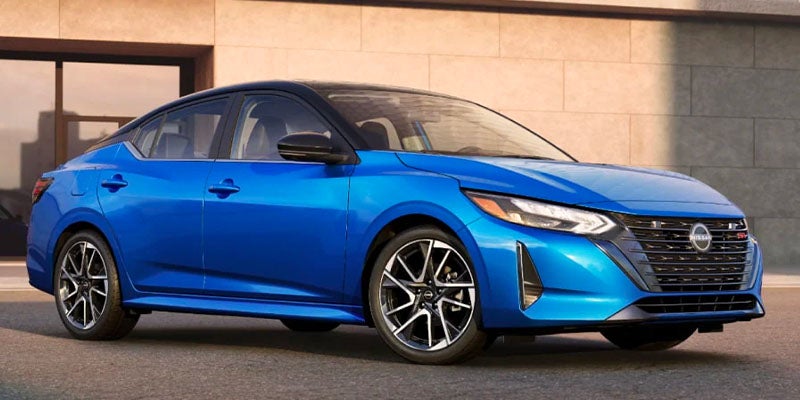 A bright blue Nissan Sentra, with a black roof and black wheels, parked in front of a modern building.