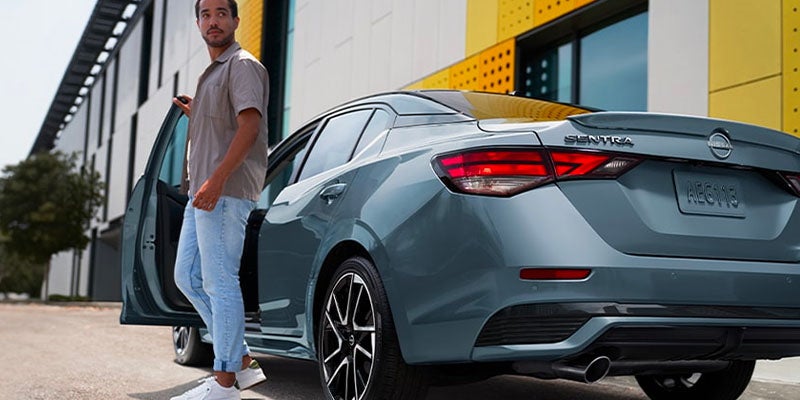 Man in casual clothes getting into a gray sedan with black wheels, parked next to a yellow and gray building.