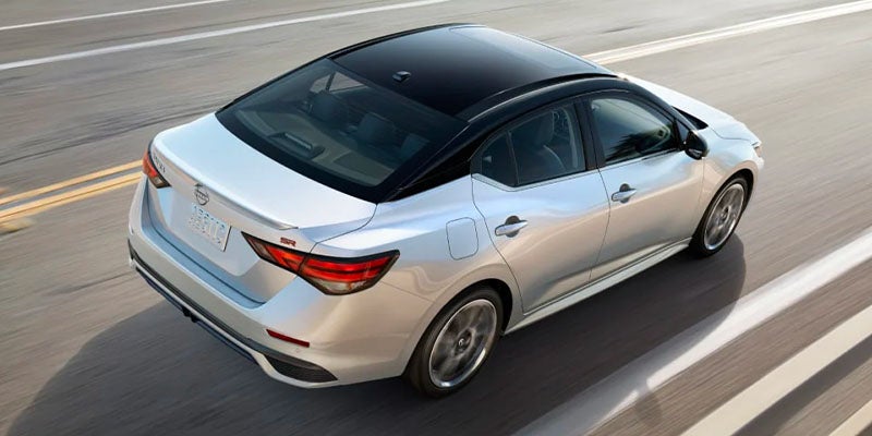 Silver Nissan sedan with a black roof drives on a highway, viewed from above and behind.